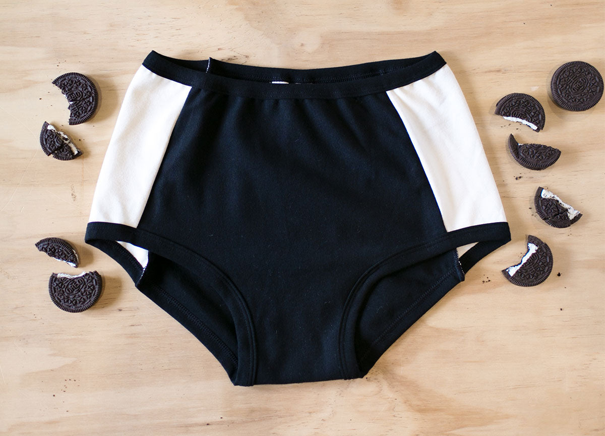 Flat lay on a wood surface and oreos around of Original style Panel Pants underwear in Cookies and Cream: black front and back with vanilla sides.