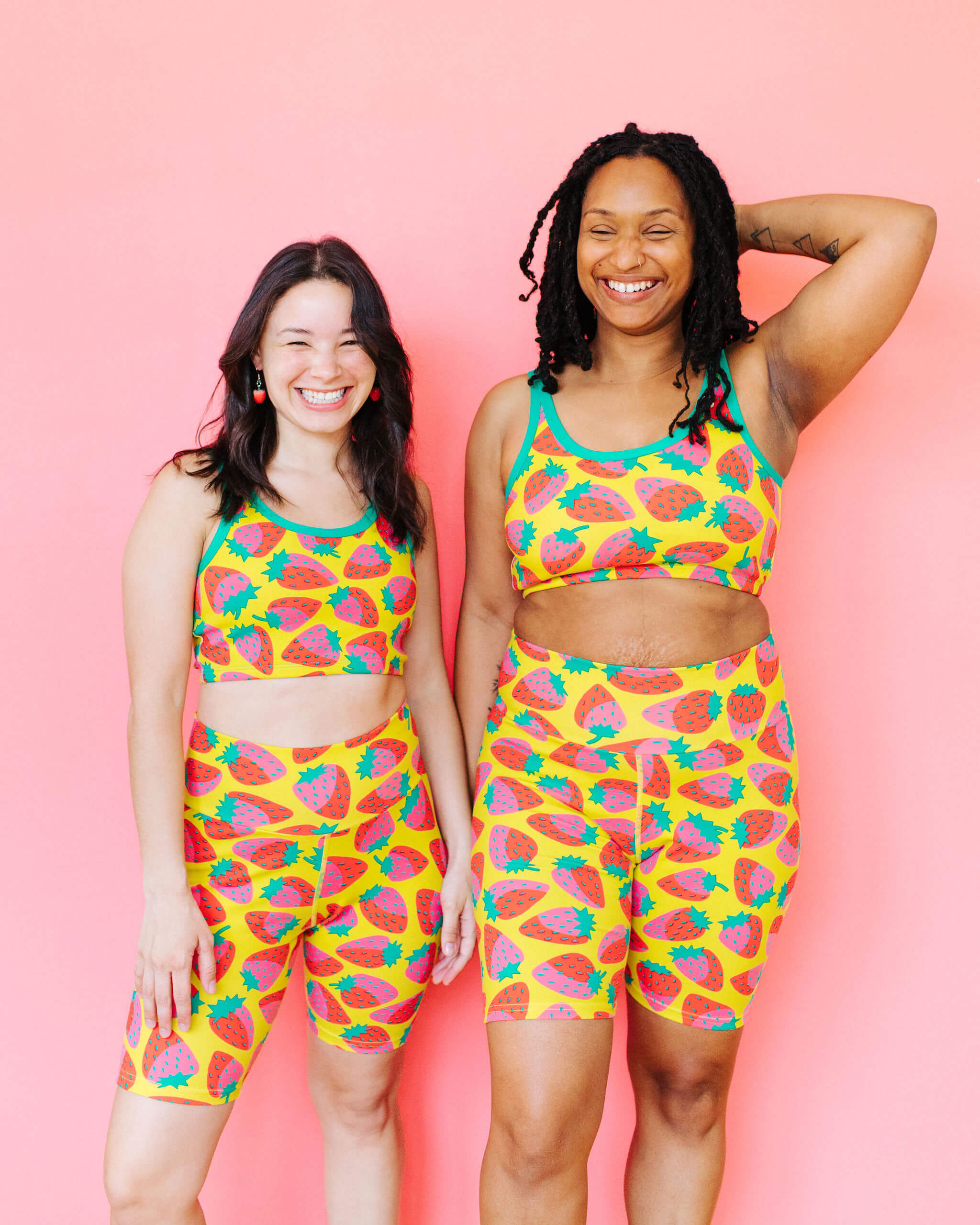 Models wearing Thunderpants Longline Bra and Bike Shorts in a Strawberry print - pink, red, and peacock green on chartreuse. 