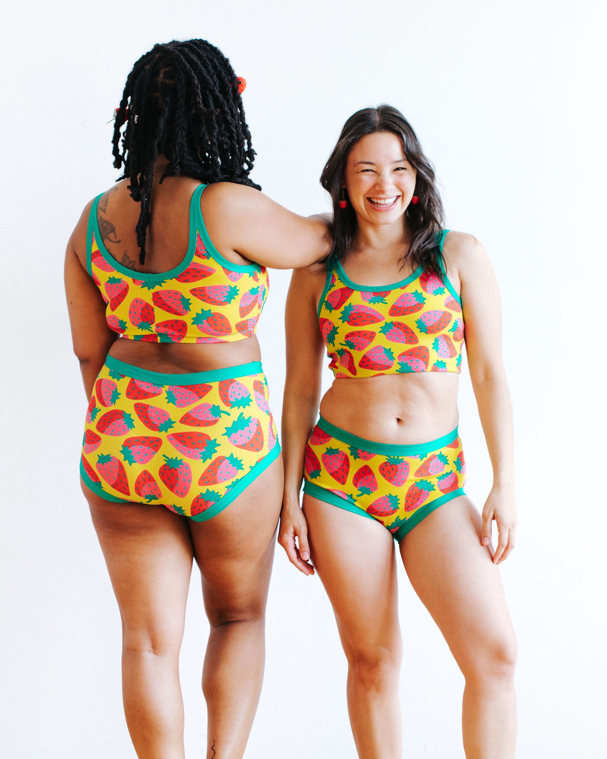Models wearing Thunderpants Longline Bra and Original style underwear in a Strawberry print - pink, red, and peacock green on chartreuse. 