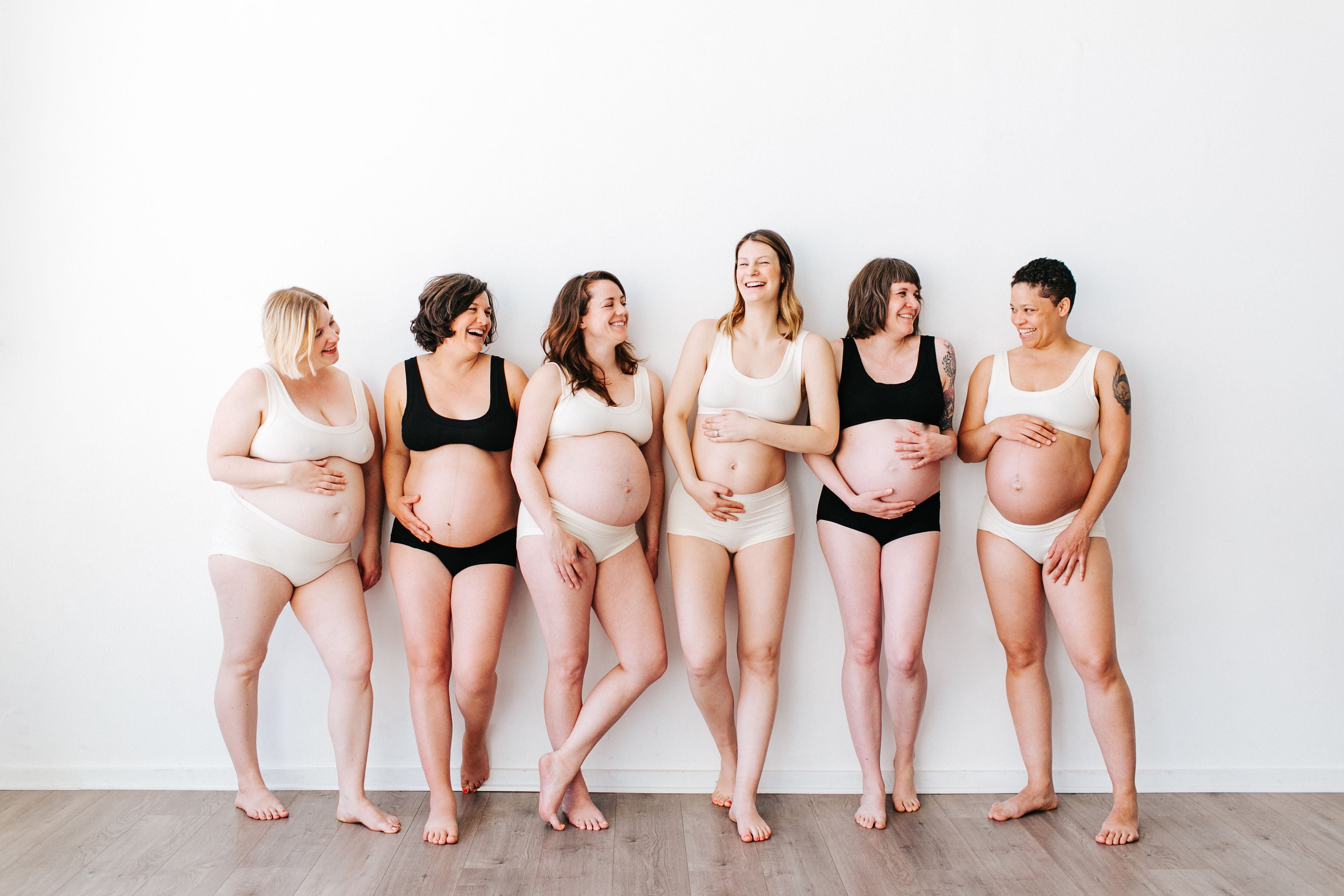 Six pregnant models standing and laughing together wearing Thunderpants Organic Cotton Original and Hipster underwear with Bralettes in both plain Black and Vanilla colors.