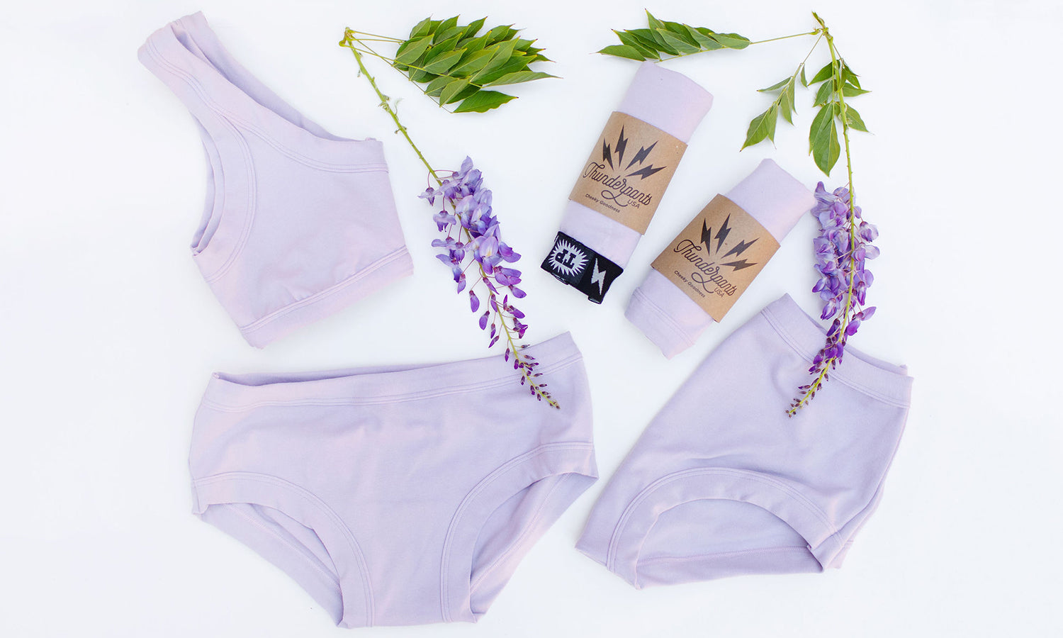 Various Thunderpants styles on a white background with wisteria plants in the color Wisterial Girl - a pruple/grey. 