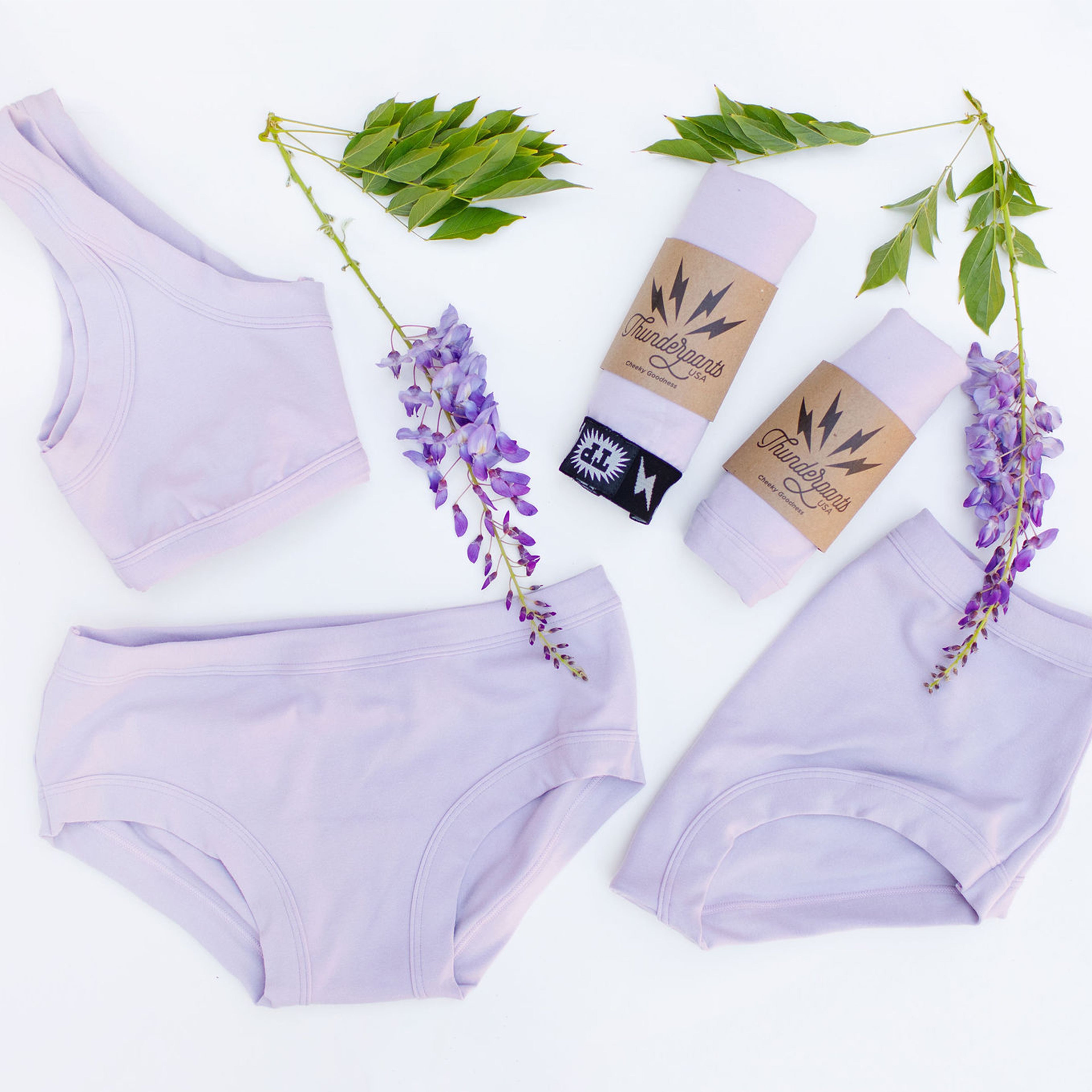 Various Thunderpants styles on a white background with wisteria plants in the color Wisterial Girl - a pruple/grey. 